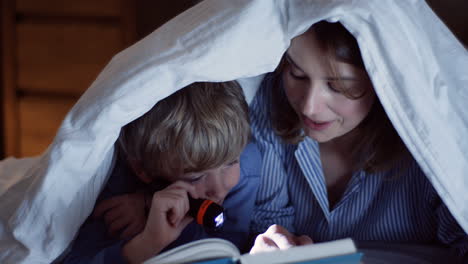 Cute-small-boy-and-his-pretty-mother-with-a-little-lantern-reading-an-interesting-book-under-the-blanket-at-night.-Close-up.-Inside.